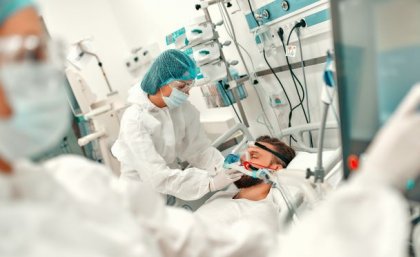 A person in a medical gown, mask and hair covering bends over a man in a bed surrounded by medical equipment.